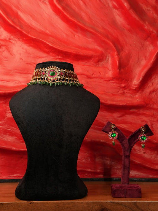 An ornate gold-toned choker necklace with green and red stones is displayed on a black mannequin bust, with matching drop earrings on a small stand against a textured red background.