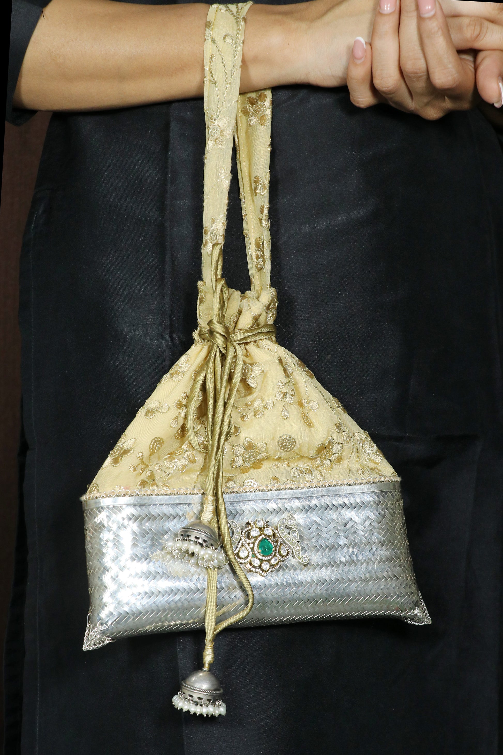 A close-up of a person holding a small drawstring pouch bag. The bag has a beige and gold patterned upper half, a metallic silver patterned lower half, and a decorative silver clasp with a green stone.