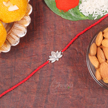 A silver leaf-shaped rakhi bracelet on a red braided cord, displayed on a textured brown surface with festive sweets, rice, almonds, and a green leaf in the background.