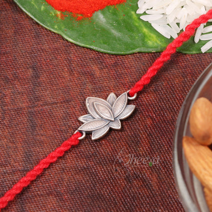 A red braided string bracelet with a silver-toned lotus flower charm, displayed on a textured brown surface with decorative elements like a green leaf, white rice, and almonds.