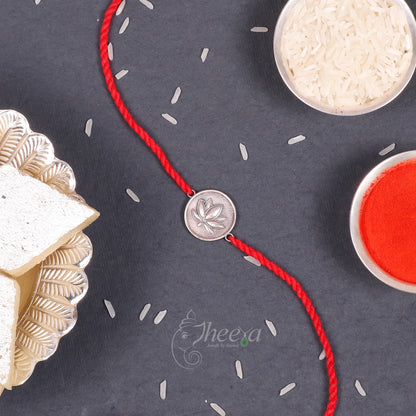 A red braided string bracelet with a silver coin charm featuring a leaf design, styled on a dark slate background with rice, red powder, and a gold-toned plate.