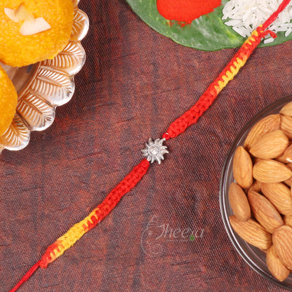 A red and yellow braided Rakhi with a silver sun charm, laid diagonally on a dark brown textured surface surrounded by festive food props like laddoo, almonds, and rice.
