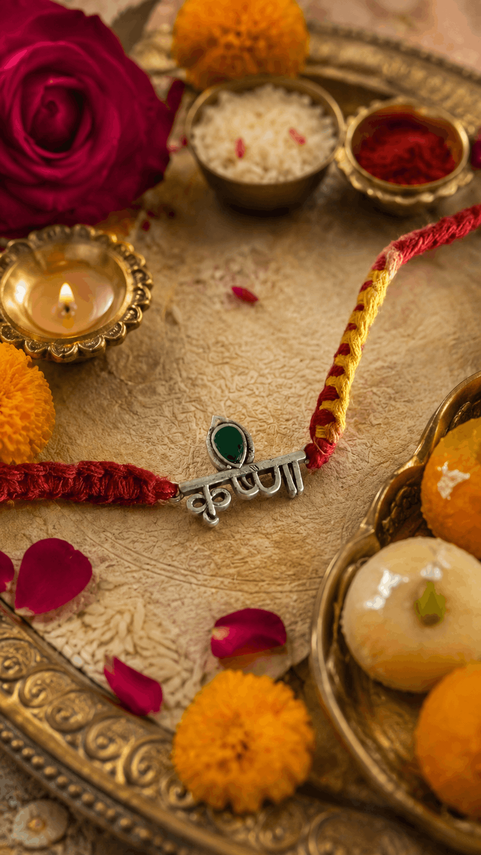 A close-up styled arrangement featuring a silver-toned Rakhi with a green teardrop-shaped stone and a Hindi nameplate, threaded with red and yellow cords on a decorative tray. The scene includes a lit diya, marigold flowers, rose petals, and sweets, with no person or animal present.