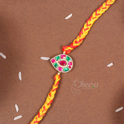 A braided yellow and red cord Rakhi bracelet with a central silver-toned evil eye charm embellished with pink and green stones, laid diagonally on a brown textured background.