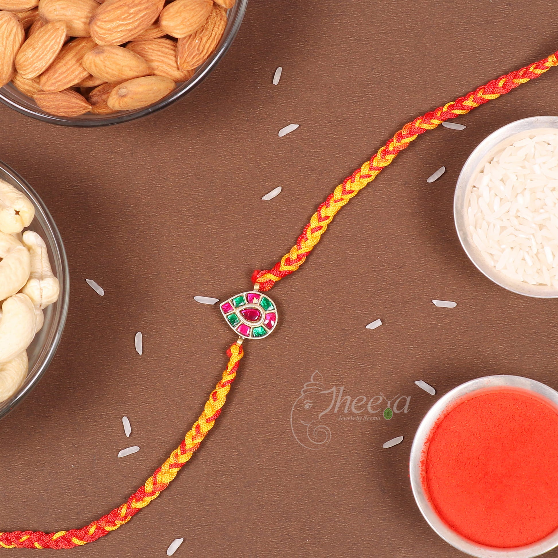 A braided Rakhi with a central decorative charm featuring pink, green, and red stones, laid out on a brown surface with bowls of almonds, rice, and red powder nearby.
