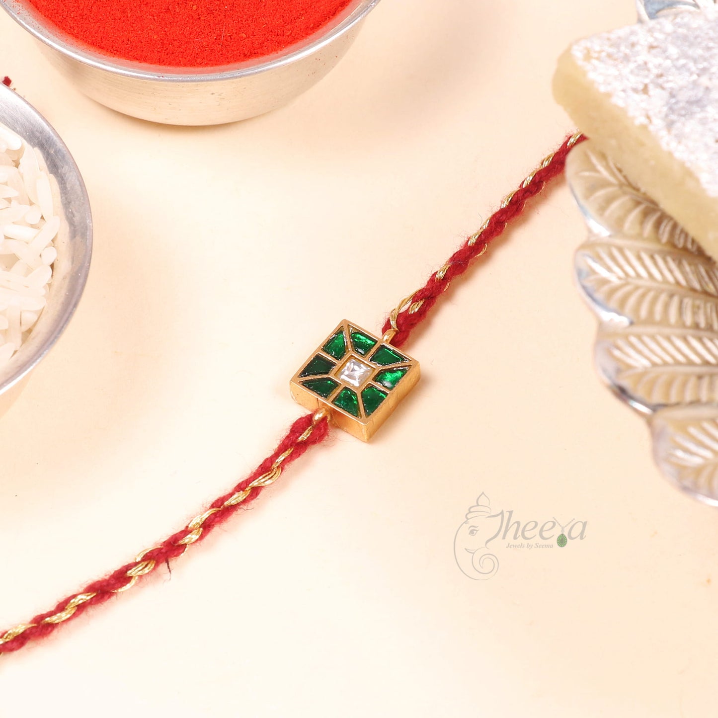 A red braided Rakhi bracelet with a central gold square charm featuring green and white inlay, displayed on a light cream surface with festive props like a bowl of red powder, rice, and a piece of sugar.