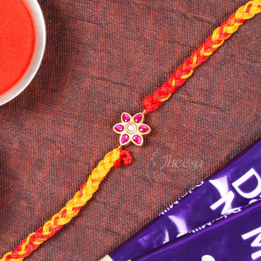 A braided Rakhi bracelet with a gold-tone flower charm set with pink stones, laid diagonally on a textured dark brown surface with a red powder dish and chocolate bar partially visible.