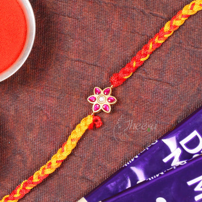 A braided Rakhi bracelet with a gold-tone flower charm set with pink stones, laid diagonally on a textured dark brown surface with a red powder dish and chocolate bar partially visible.