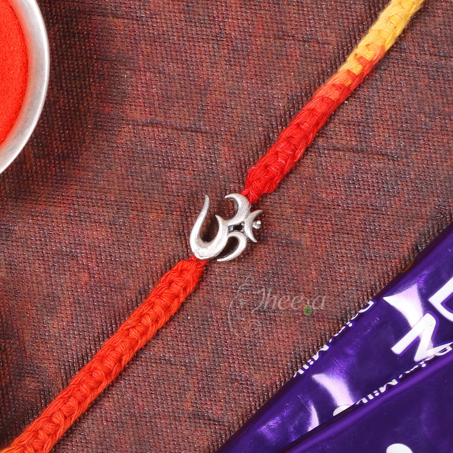 A braided Rakhi featuring a red and yellow gradient cord with a silver Om symbol charm, laid diagonally on a textured brown fabric background with a bowl of red powder and a purple packet partially visible.