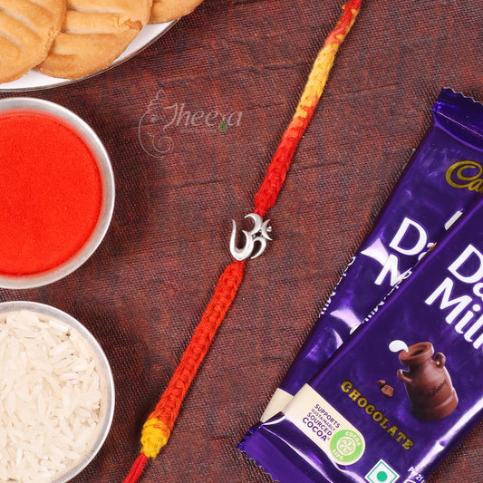 A red and orange braided Rakhi with a silver Om symbol charm, styled on a dark brown textured surface alongside a plate of cookies, a bowl of red powder, a bowl of white rice, and two chocolate milk packets.