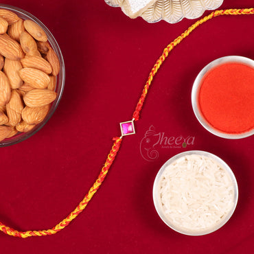 A braided yellow and red Rakhi with a small pink stone charm, laid out on a deep red fabric background alongside traditional festive props including a bowl of almonds, a bowl of white rice, a bowl of red powder, and a silver shell.
