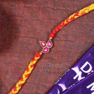 A braided Rakhi bracelet with red and yellow threads and a gold-toned butterfly charm with pink stones, laid diagonally on a textured brown fabric next to a purple chocolate bar.