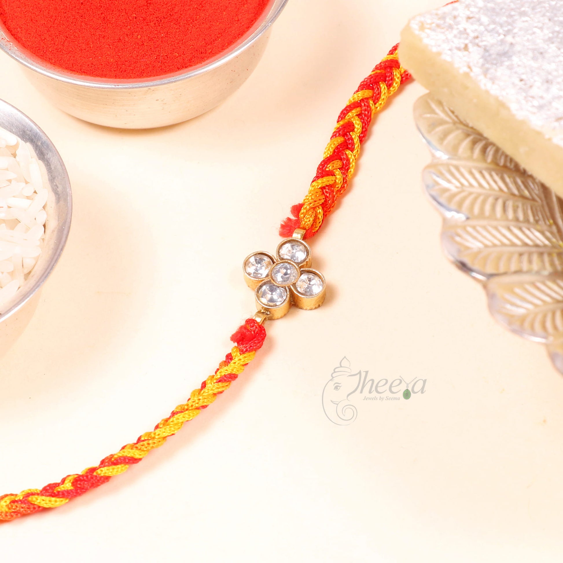 A braided Rakhi bracelet in red and yellow thread with a gold-toned flower charm set with clear stones, displayed on a light cream surface with festive props including a bowl of red powder, rice, and a block of soap.