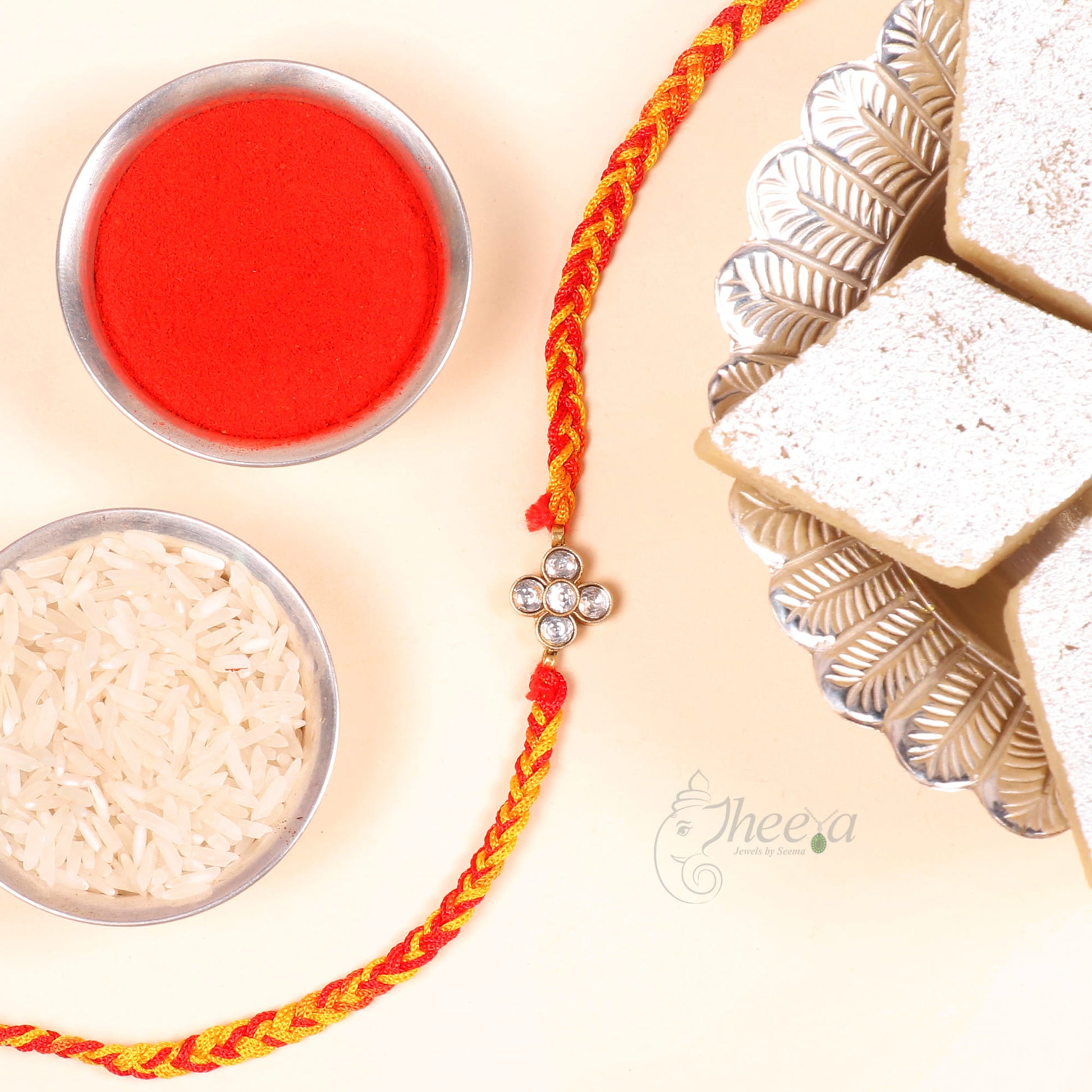 A braided Rakhi with red and yellow threads and a silver-toned charm, arranged on a cream background with bowls of red powder and rice, and white sweets on the right.